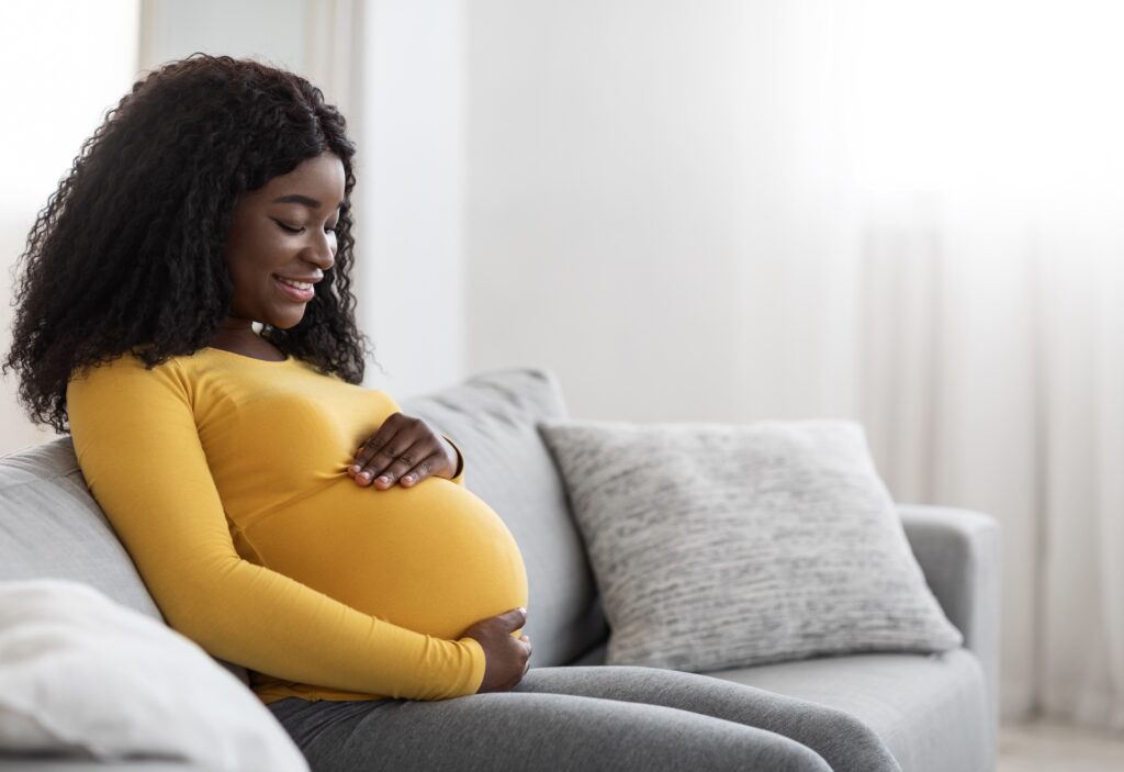 Joyful black pregnant woman hugging her big tummy, communicating with her baby, sitting on sofa at living room.