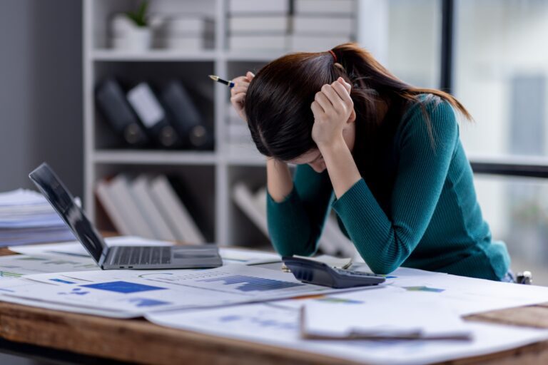 A woman has her head in her hands while sat at her desk in front of the computer. Her tiredness may be related to anemia or iron deficiency