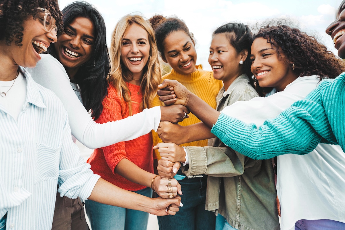 Diverse group of women of different ethnicities and ages putting their hands together in a circle, showing unity and support.