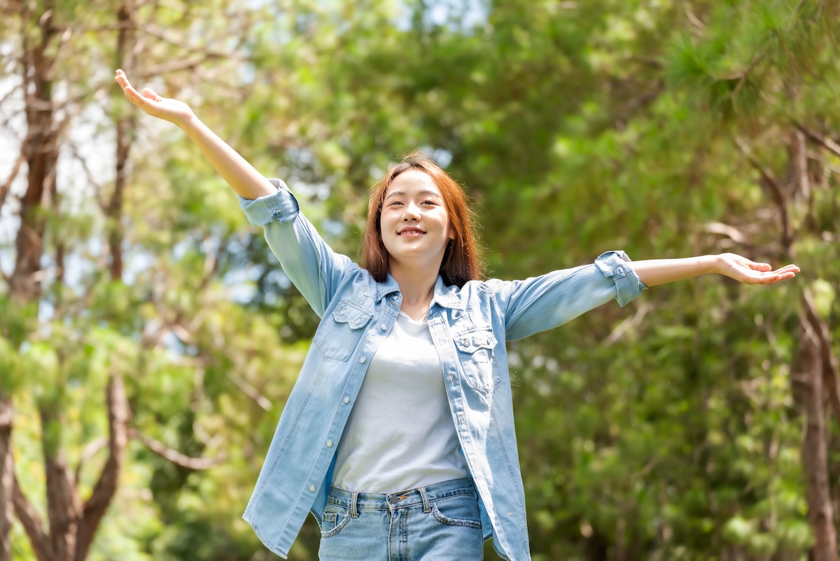 Woman with arms raised joyfully outdoors in nature, smiling and expressing freedom and wellness.